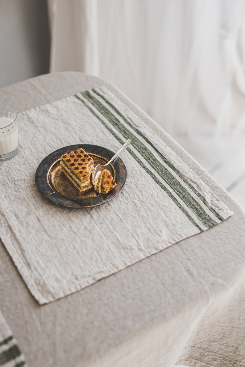 May include: A piece of layered cake with a brown and white filling on a silver plate with a spoon. The plate is on a beige linen tablecloth with green stripes.