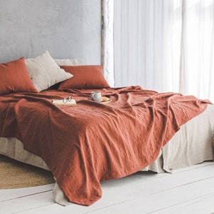 Pu&ograve; includere: Un copriletto in lino color ruggine con federe abbinate, una tazza in ceramica bianca e un libro sopra il letto. Il letto &egrave; fatto con un lenzuolo in lino bianco e ha un tappeto in fibra naturale sotto.