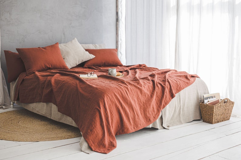 Pu&ograve; includere: Un copriletto in lino color ruggine con federe abbinate, una tazza in ceramica bianca e un libro sopra il letto. Il letto &egrave; fatto con un lenzuolo in lino bianco e ha un tappeto in fibra naturale sotto.
