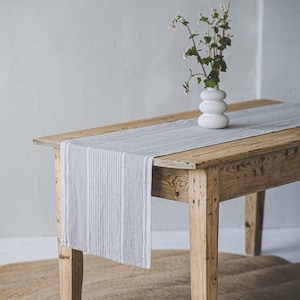 May include: A rustic wooden table with a gray and white striped linen table runner. A white ceramic vase with a small bouquet of white flowers sits on the table runner.