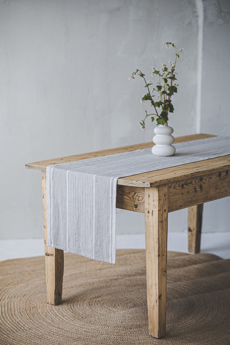 May include: A rustic wooden table with a gray and white striped linen table runner. A white ceramic vase with a small bouquet of white flowers sits on the table runner.