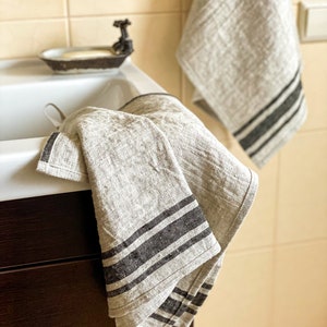 Puede incluir: Dos toallas de cocina de lino grises con rayas negras colgadas en un ba&ntilde;o. Las toallas est&aacute;n dobladas y colgadas sobre un armario.