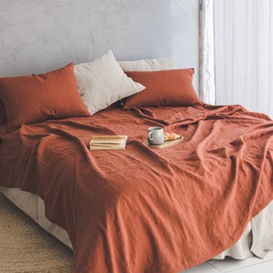 Pu&ograve; includere: Un set di biancheria da letto in lino color ruggine con due cuscini e un vassoio con una tazza e un dolce su un letto con un tappeto in fibra naturale.