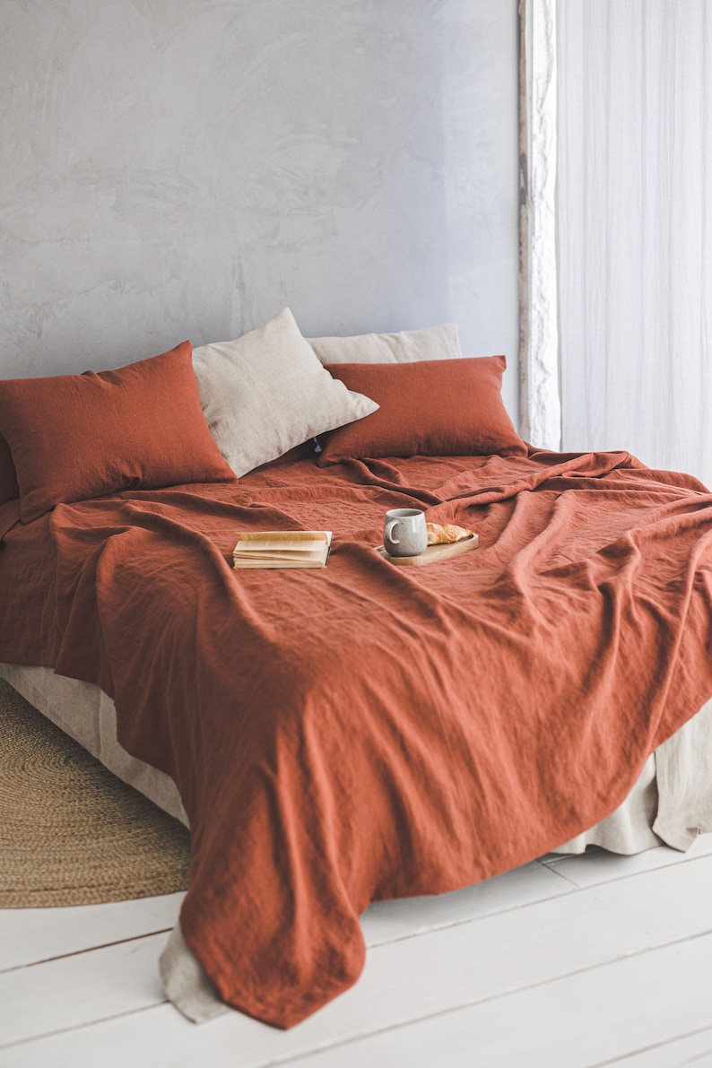 Pu&ograve; includere: Un set di biancheria da letto in lino color ruggine con due cuscini e un vassoio con una tazza e un dolce su un letto con un tappeto in fibra naturale.