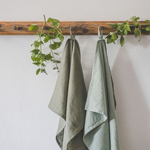 May include: Two rectangular kitchen towels hanging from a wooden wall rack. One towel is a deep olive green, and the other is a lighter sage green. Both towels feature a small fabric loop at the top for hanging and have a visible woven texture.