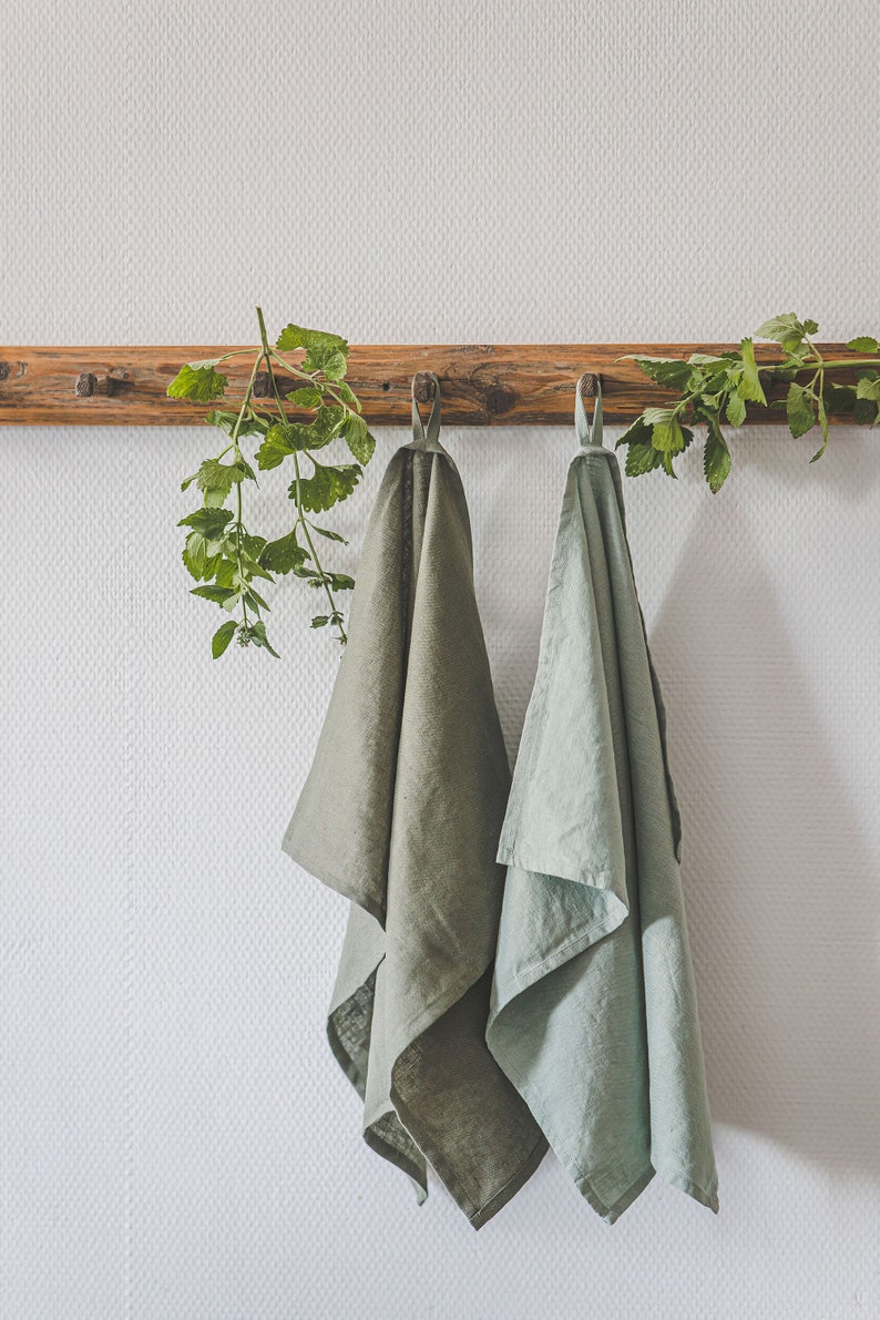 May include: Two rectangular kitchen towels hanging from a wooden wall rack. One towel is a deep olive green, and the other is a lighter sage green. Both towels feature a small fabric loop at the top for hanging and have a visible woven texture.