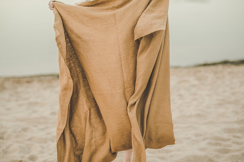 May include: A brown linen throw blanket is held up in front of a sandy beach background.