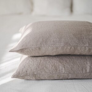 May include: Two brown linen pillowcases stacked on a white bed. The pillowcases are made of a soft, natural fabric and have a subtle texture.