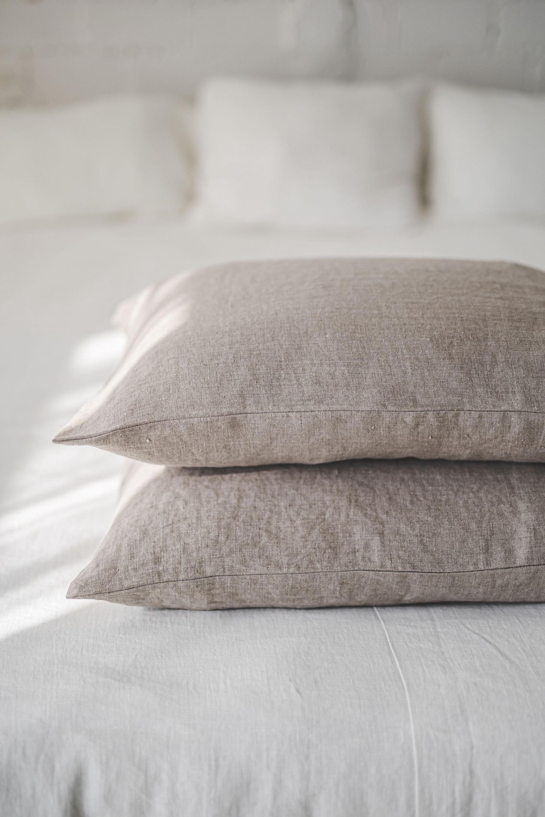 May include: Two brown linen pillowcases stacked on a white bed. The pillowcases are made of a soft, natural fabric and have a subtle texture.