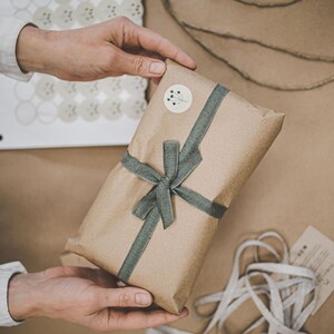 May include: A rectangular gift wrapped in brown kraft paper, tied with a sage green fabric ribbon in a bow. A small circular sticker featuring a black line drawing of a sprig is affixed to the top. The package is held against a neutral background.