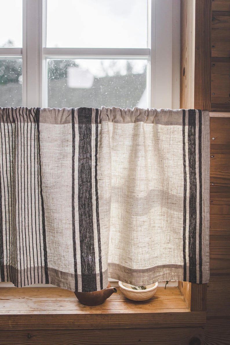 May include: A window with a white frame and a view of a cloudy sky. A white linen curtain with black stripes hangs in front of the window. A small brown teapot and a white bowl with green leaves are on the windowsill.
