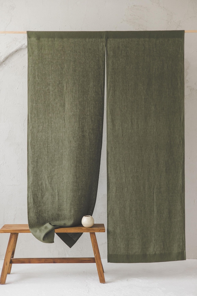 May include: Two olive green linen curtains hanging from a wooden rod. The curtains are textured and have a subtle sheen. A small wooden bench with a white ceramic bowl sits in front of the curtains.