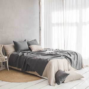Pu&ograve; includere: Un letto in lino grigio con una coperta in lino beige e cuscini grigi in una camera da letto con pareti bianche e tende bianche trasparenti.