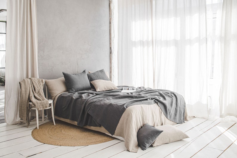 Pu&ograve; includere: Un letto in lino grigio con una coperta in lino beige e cuscini grigi in una camera da letto con pareti bianche e tende bianche trasparenti.