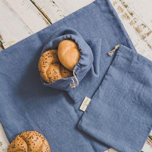 Puede incluir: Una bolsa de pan de lino azul con cierre de cordón, un saco a juego y un mantel individual. Dos panes están dentro de la bolsa. Un pan con semillas está en el mantel individual. Los artículos están sobre una superficie de madera blanca.