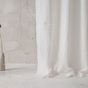 May include: A long white curtain with a textured fabric surface hangs vertically. Beside the curtain, a small, light gray, irregularly shaped ceramic vase holds a single dried brown poppy seed head on a thin, slender stem.