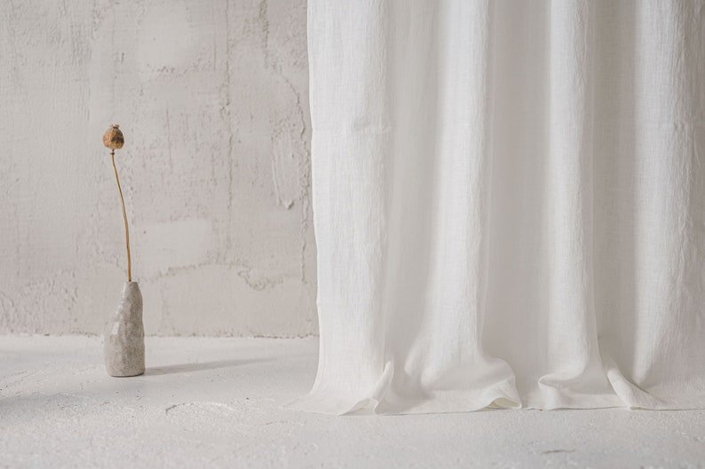 May include: A long white curtain with a textured fabric surface hangs vertically. Beside the curtain, a small, light gray, irregularly shaped ceramic vase holds a single dried brown poppy seed head on a thin, slender stem.