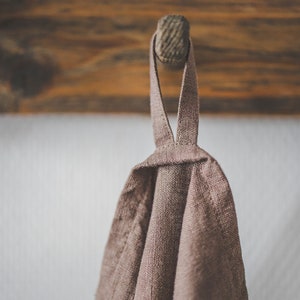 May include: A taupe-colored linen kitchen towel featuring a simple fabric loop at the top for hanging. The textile has a visible woven texture and is draped downwards, showcasing the natural, earthy tone of the fabric in a neutral light.