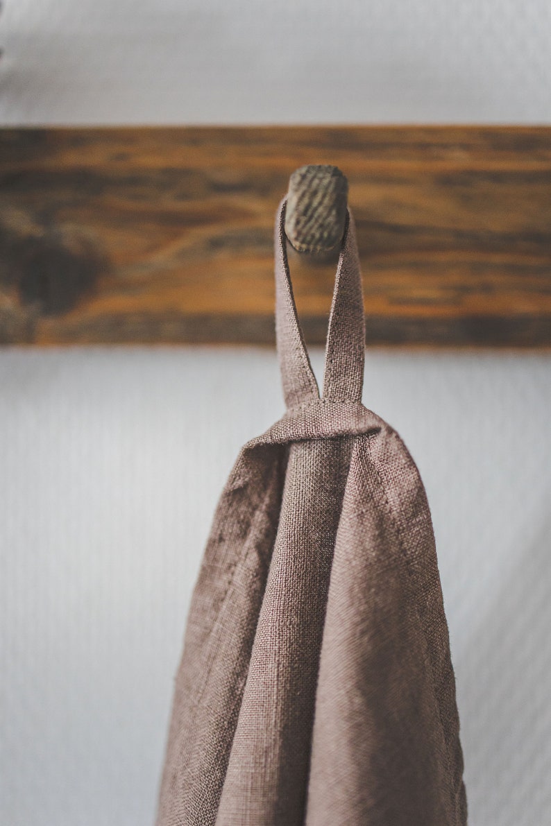 May include: A taupe-colored linen kitchen towel featuring a simple fabric loop at the top for hanging. The textile has a visible woven texture and is draped downwards, showcasing the natural, earthy tone of the fabric in a neutral light.