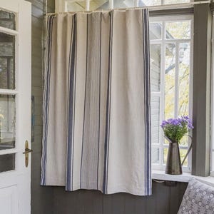 Può includere: Tenda beige a righe verticali blu davanti a una finestra. La tenda è realizzata in tessuto chiaro con strisce blu verticali. Una porta bianca è a sinistra. Un vaso di metallo con fiori viola è sul davanzale.