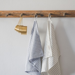May include: A set of gold-toned metal measuring cups hanging alongside two kitchen towels. One towel is light blue with thin dark stripes, and the other is off-white with thin dark stripes. Both towels feature a small fabric loop for hanging.