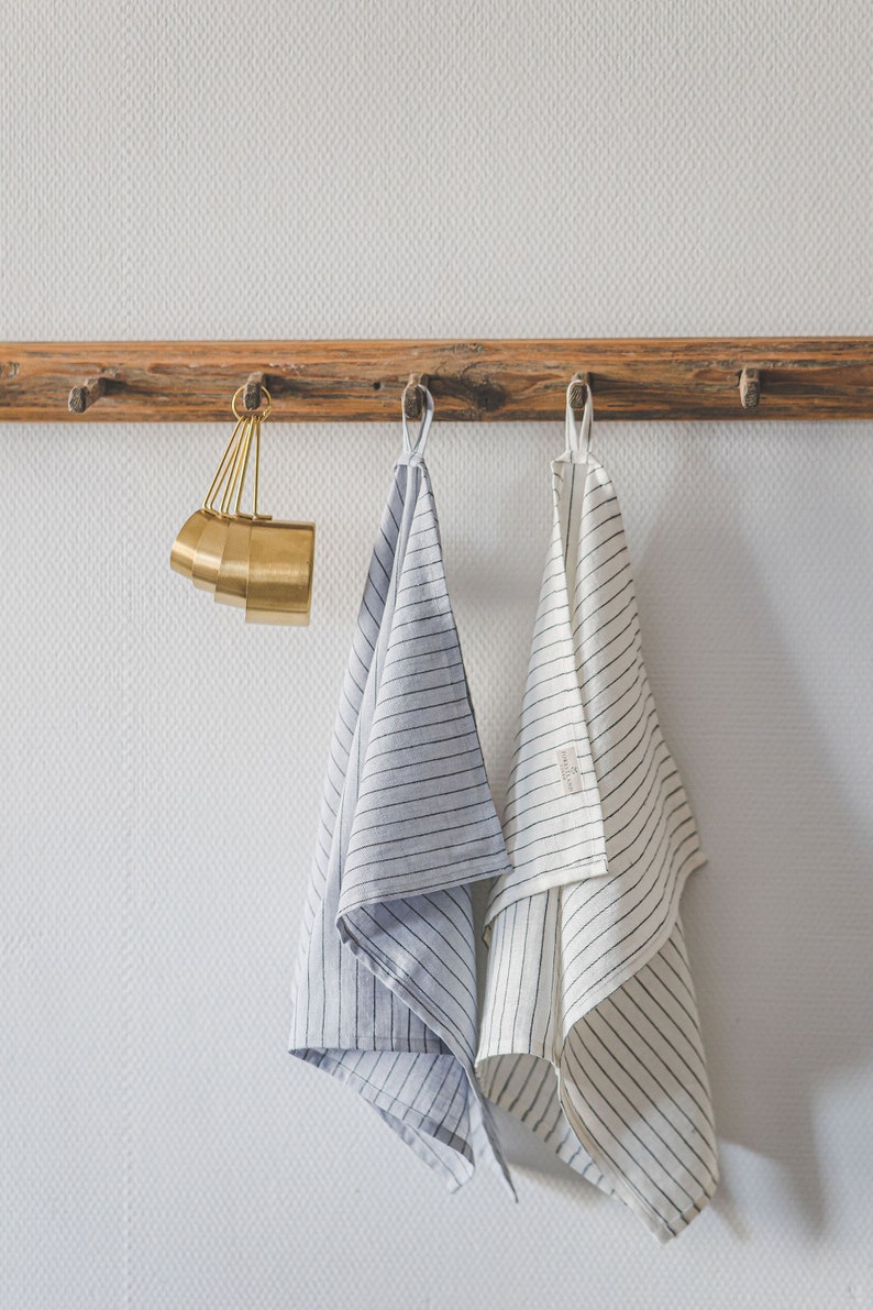 May include: A set of gold-toned metal measuring cups hanging alongside two kitchen towels. One towel is light blue with thin dark stripes, and the other is off-white with thin dark stripes. Both towels feature a small fabric loop for hanging.