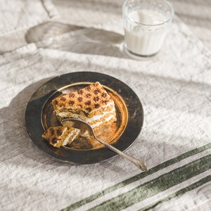 May include: A slice of layered cake with a brown and white frosting on a dark brown plate with a silver spoon. The plate is on a light brown linen tablecloth with green stripes.