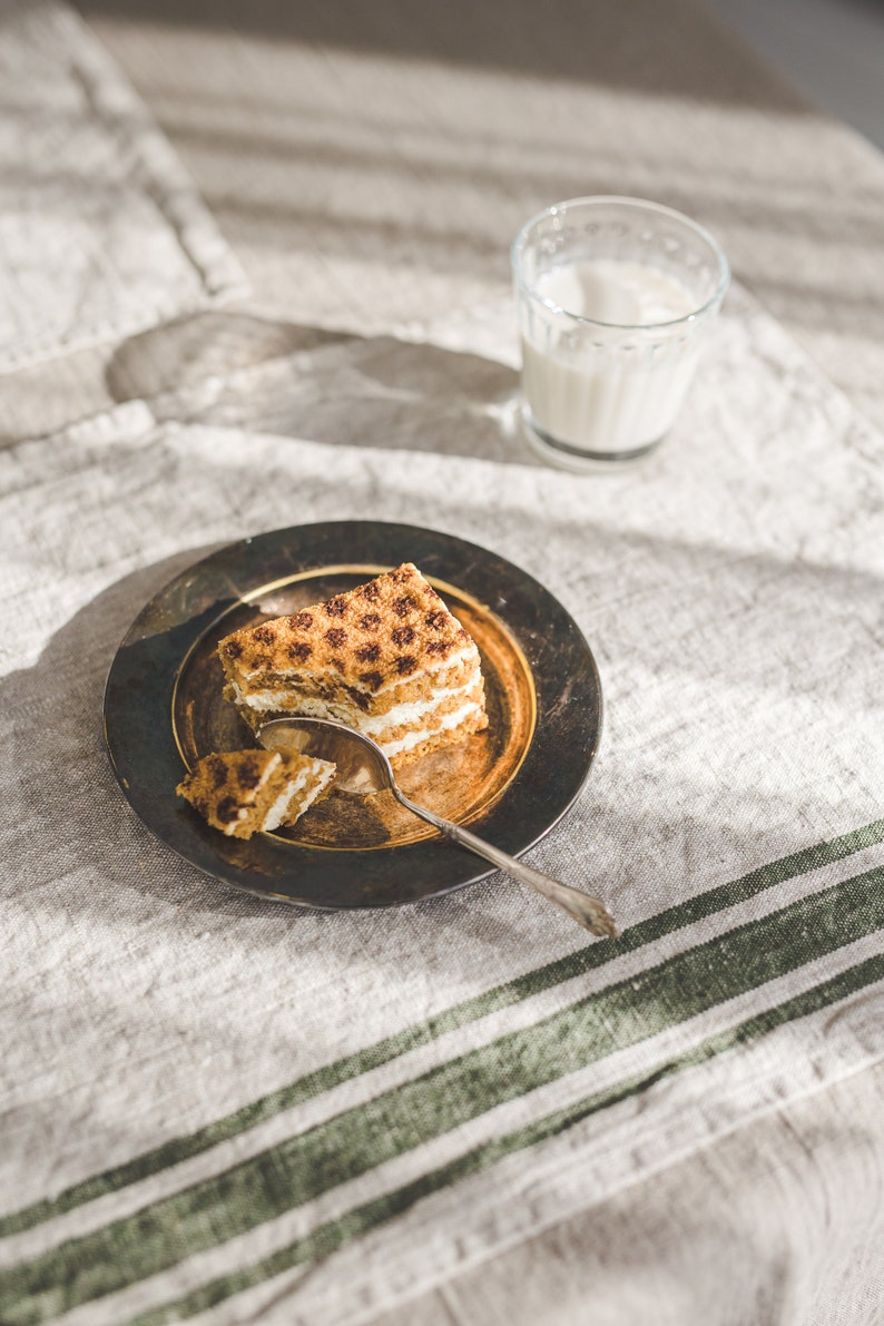 May include: A slice of layered cake with a brown and white frosting on a dark brown plate with a silver spoon. The plate is on a light brown linen tablecloth with green stripes.