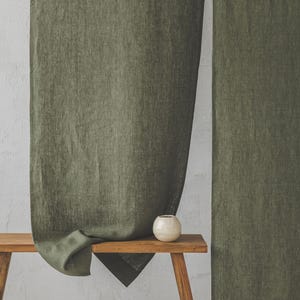 May include: Two olive green linen curtains hanging on a white wall. A small wooden bench with a white ceramic vase sits in front of the curtains.