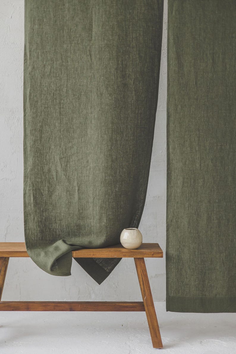 May include: Two olive green linen curtains hanging on a white wall. A small wooden bench with a white ceramic vase sits in front of the curtains.