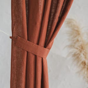 Op de afbeelding: Een roestkleurig gordijn met een zichtbare geweven textuur en een bijpassende stoffen embrasse die de stof bijeenhoudt. Het gordijn hangt in verticale plooien en vertoont een diepe terracottakleur en een semi-transparante kwaliteit bij lichtinval.