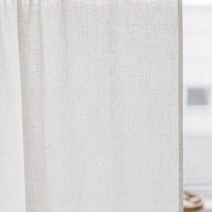 May include: A semi-sheer off-white curtain panel with a visible woven texture, featuring a rod pocket header at the top. The fabric is light-colored and hangs vertically from a horizontal rod, displaying a natural weave pattern throughout the material.