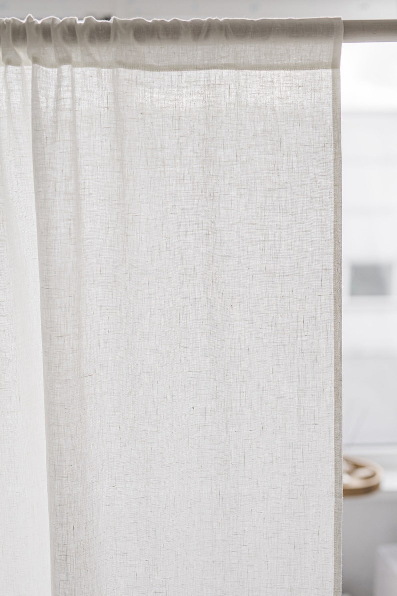 May include: A semi-sheer off-white curtain panel with a visible woven texture, featuring a rod pocket header at the top. The fabric is light-colored and hangs vertically from a horizontal rod, displaying a natural weave pattern throughout the material.
