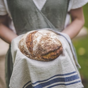 Puede incluir: Una persona sosteniendo un pan de masa madre reci&eacute;n horneado envuelto en una toalla de cocina de lino blanco con rayas azules.