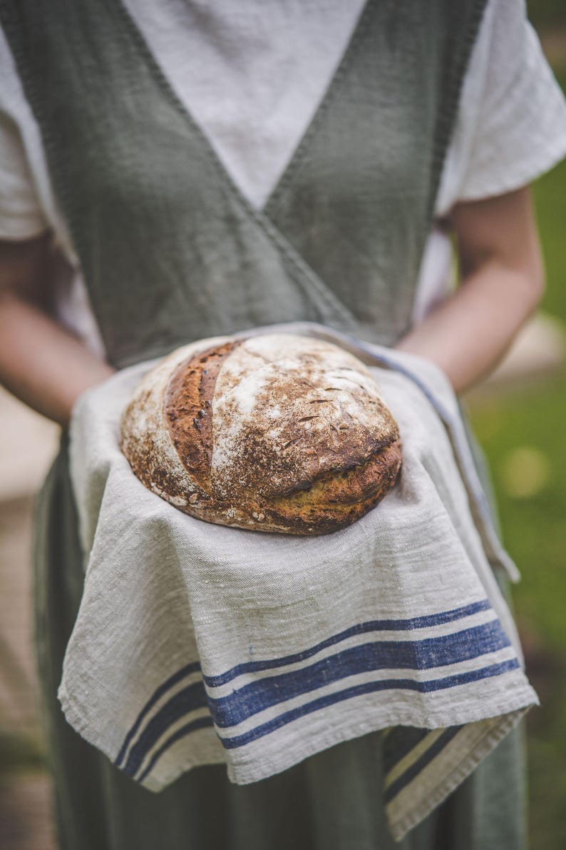 Puede incluir: Una persona sosteniendo un pan de masa madre reci&eacute;n horneado envuelto en una toalla de cocina de lino blanco con rayas azules.