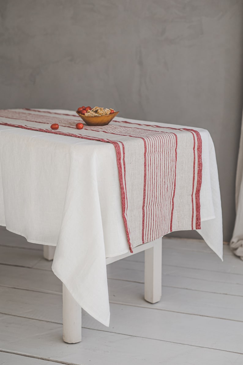 May include: White linen tablecloth with a red and white striped runner. The runner is centered on the table and has a bowl of red berries on top.