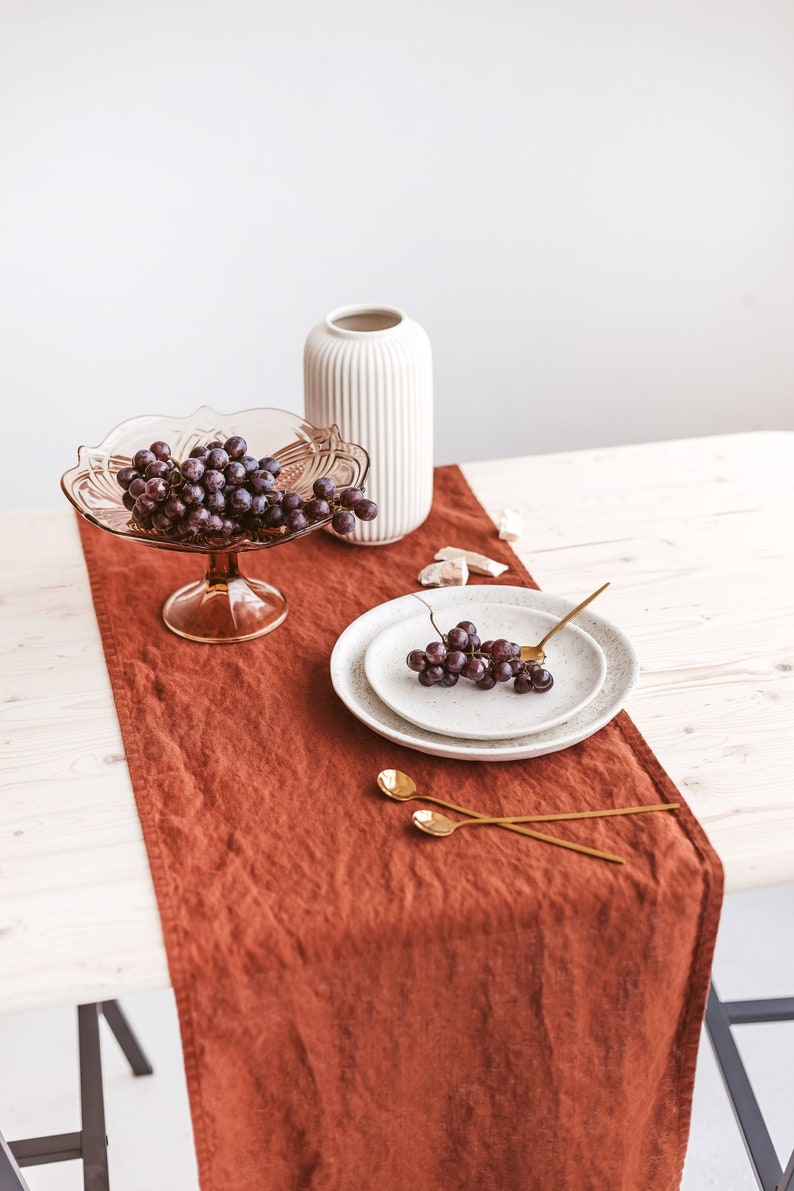 May include: A brown linen table runner with a white ceramic vase, a glass bowl of grapes, two gold spoons, and a white speckled plate with grapes on a white wooden table.