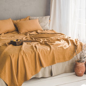 Pu&ograve; includere: Un set di biancheria da letto in lino giallo senape con due federe e un copripiumino. La biancheria da letto &egrave; disposta su un letto con una testata grigio chiaro e un pavimento in legno bianco. Il letto si trova in una camera da letto con una finestra sullo sfondo.