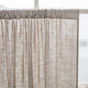May include: A light beige, semi-sheer curtain panel with a visible woven texture. The curtain features a rod pocket header at the top, creating soft, vertical gathers as it hangs from a white curtain rod, allowing light to filter through the fabric.
