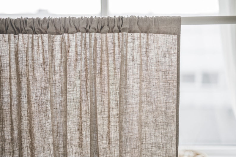 May include: A light beige, semi-sheer curtain panel with a visible woven texture. The curtain features a rod pocket header at the top, creating soft, vertical gathers as it hangs from a white curtain rod, allowing light to filter through the fabric.