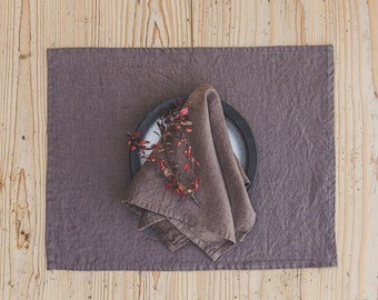 May include: A rectangular dark brown linen placemat featuring a matching folded napkin placed on a round plate. A small branch with red berries and autumn-colored leaves rests on top of the napkin, adding a natural accent to the table setting.