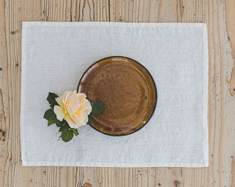 Puede incluir: Un plato redondo de cerámica marrón con un borde oscuro, que presenta una sola rosa de color amarillo pálido con hojas verdes descansando en su borde, todo centrado en un mantel individual rectangular de lino blanco con un dobladillo visible.