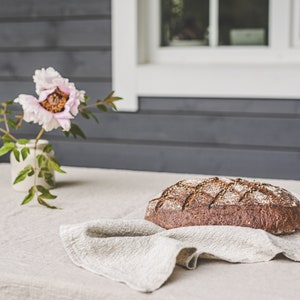 May include: A loaf of freshly baked bread sits on a linen tablecloth with a pink flower in a vase in the background.