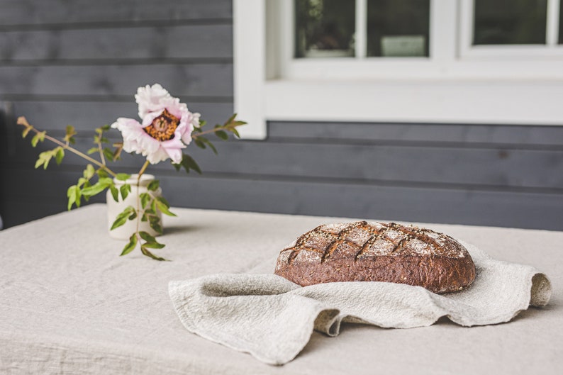 May include: A loaf of freshly baked bread sits on a linen tablecloth with a pink flower in a vase in the background.