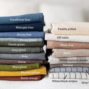 Pode incluir: Duas pilhas de tecidos dobrados em várias cores e padrões. Pilha esquerda: azul centáurea, azul meia-noite, azul jeans, verde geada, verde floresta, verde cinza, cinza, cacau, verde musgo, amarelo mimosa e laranja queimado. Pilha direita: amarelo baunilha, branco off-white, rosa névoa, areia, xadrez branco/natural, natural, listras branco/preto e listras cinza/preto.
