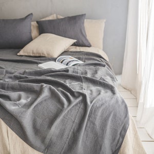 Pu&ograve; includere: Una coperta di lino grigia drappeggiata su un letto con cuscini e lenzuola di lino beige. La coperta &egrave; piegata e ha un aspetto naturale e rilassato.