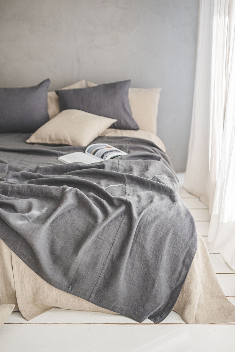 Pu&ograve; includere: Una coperta di lino grigia drappeggiata su un letto con cuscini e lenzuola di lino beige. La coperta &egrave; piegata e ha un aspetto naturale e rilassato.