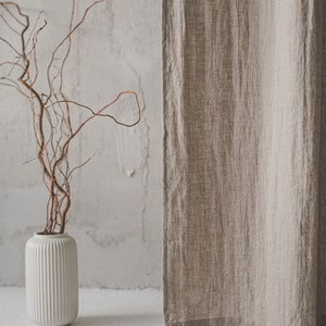 May include: A beige linen curtain hangs vertically next to a white cylindrical vase with vertical ribbing. The vase contains several thin, brown, twisted branches that extend upward. The fabric of the curtain shows a visible woven texture.