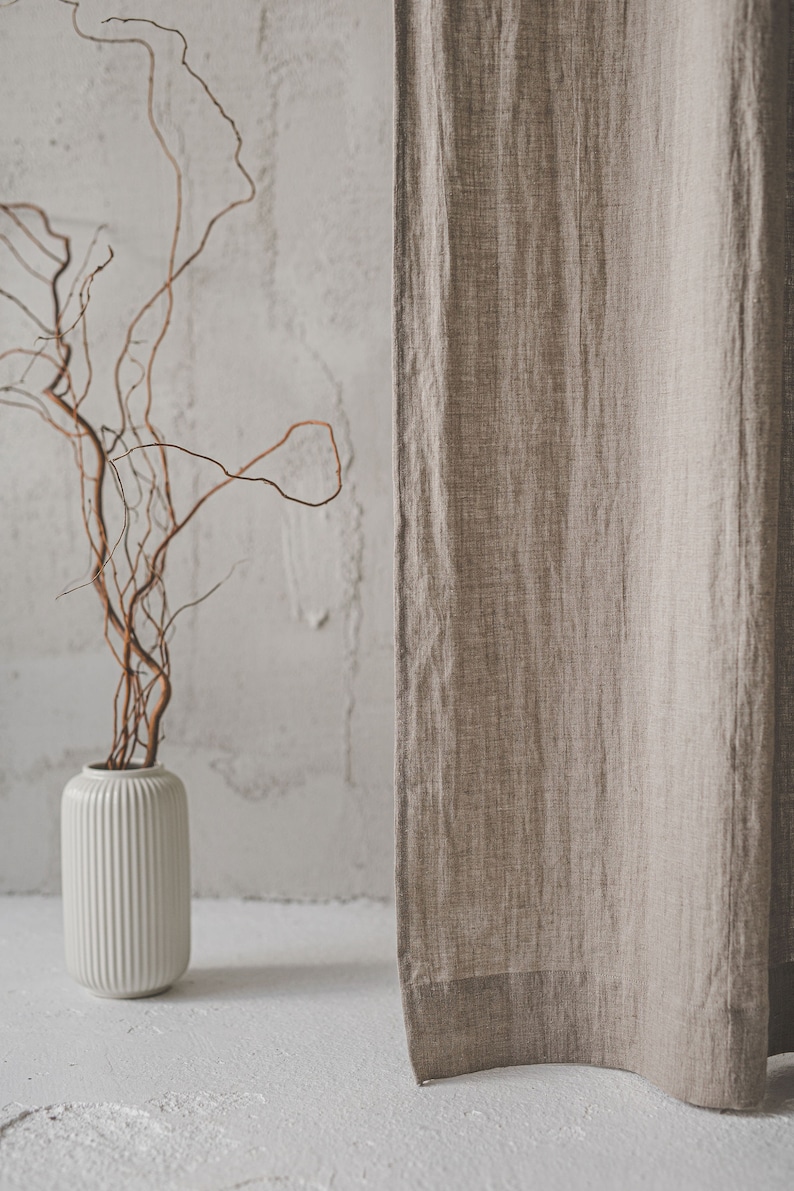May include: A beige linen curtain hangs vertically next to a white cylindrical vase with vertical ribbing. The vase contains several thin, brown, twisted branches that extend upward. The fabric of the curtain shows a visible woven texture.