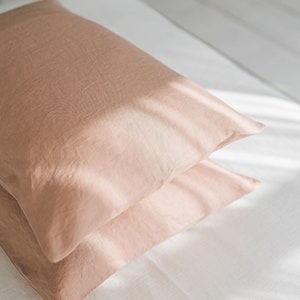 May include: Two peach-colored pillowcases stacked on a white bed sheet. The pillowcases are made of a soft, textured fabric.